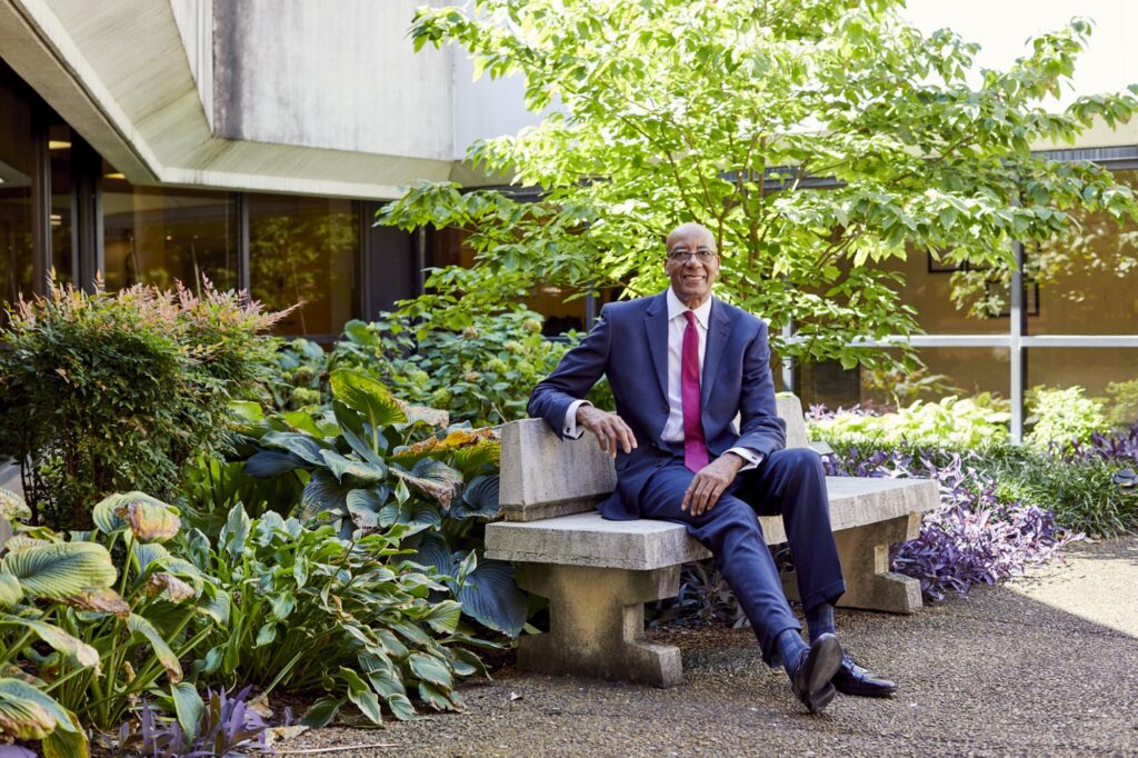 Dr. Joseph Webb, D.Sc., MSHA, FACHE, seated outdoors on a stone bench, smiling