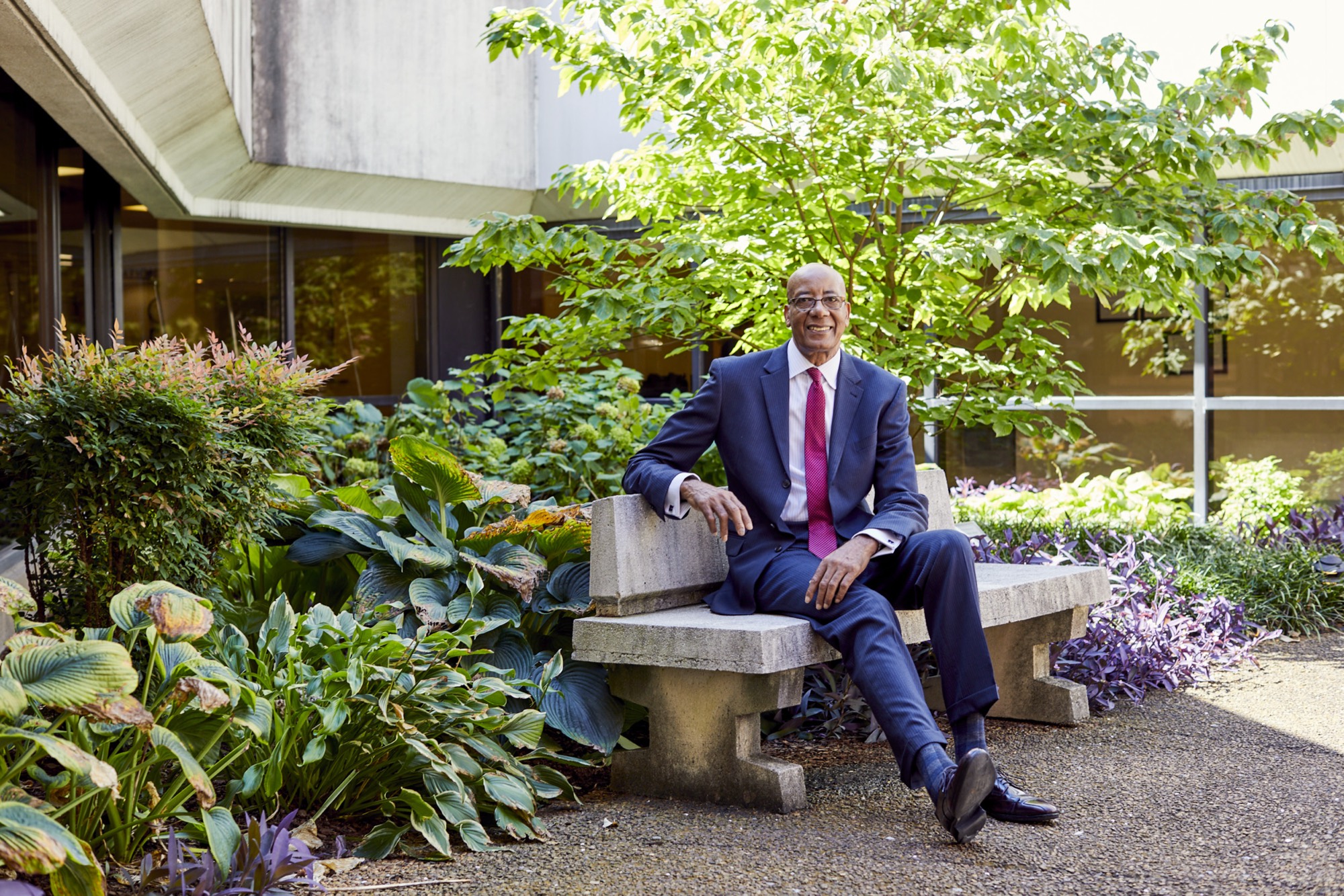 Dr. Joseph Webb, D.Sc., MSHA, FACHE, seated outdoors on a stone bench, smiling